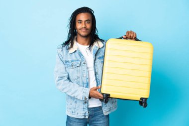 Mavi arka planda saç örgüleri olan genç Afrikalı Amerikalı bir adam seyahat çantasıyla tatilde.