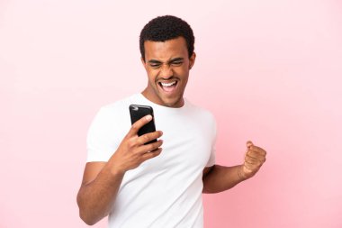 İzole edilmiş pembe arka planda cep telefonu kullanan ve zafer jesti yapan Afro-Amerikalı yakışıklı bir adam.
