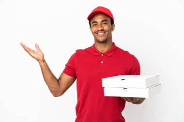 Afro-Amerikalı pizza teslimatçısı izole edilmiş beyaz duvardan pizza kutularını alıyor. Gelmesi için ellerini yana uzatıyor.