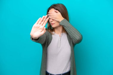 Mavi arka planda izole edilmiş genç beyaz kadın jesti durdurup yüzünü kapatıyor.