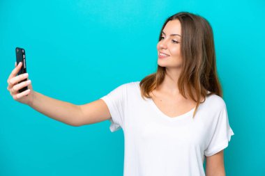 Mavi arka planda izole edilmiş beyaz bir kadın cep telefonuyla selfie çekiyor.