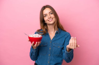 Beyaz bir kadın elinde bir kase mısır gevreği tutuyor pembe arka planda izole edilmiş bir şekilde benimle gelmesi için davet ediyor. Gelmene sevindim.