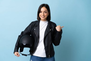 Mavi arka planda izole edilmiş motosiklet kaskı tutan kadın bir ürünü sunmak için kenarı işaret ediyor.