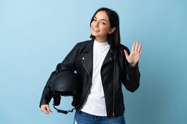 Mavi arka planda izole edilmiş motosiklet kaskı tutan kadın mutlu bir ifadeyle selamlıyor.
