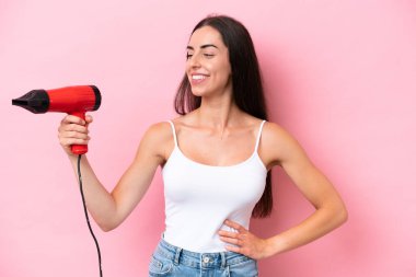 Kafkasyalı genç bir kadın elinde saç kurutma makinesiyle pembe arka planda mutlu bir ifadeyle izole edilmiş.