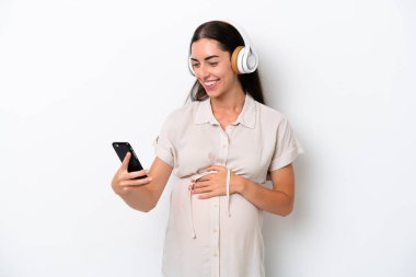 Beyaz arka planda izole edilmiş genç beyaz bir kadın hamile ve cep telefonuyla müzik dinliyor.
