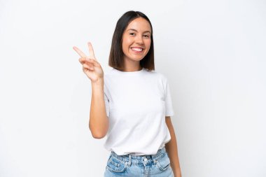 Beyaz arka planda izole edilmiş genç beyaz kadın gülümsüyor ve zafer işareti gösteriyor.