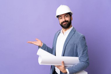Miğferli genç mimar, izole edilmiş mor arka plan üzerinde planları var. Davet etmek için ellerini yana uzatıyor.