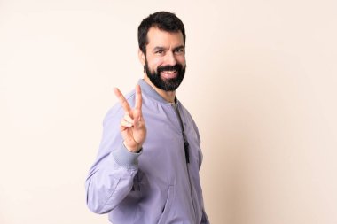Sakallı beyaz bir adam izole edilmiş arka planda ceket giyiyor gülümsüyor ve zafer işareti gösteriyor.