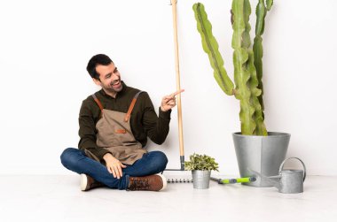 Bahçıvan içeride yerde oturuyor. Parmakla işaret ediyor ve bir ürün sunuyor.