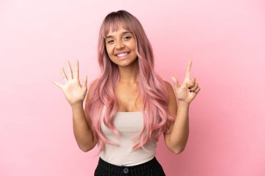 Pembe saçlı melez kadın pembe arka planda izole edilmiş yediyi parmaklarıyla sayıyor.