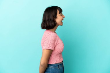 Genç melez kadın, mavi arka planda izole edilmiş. Yan pozisyonda gülüyor.