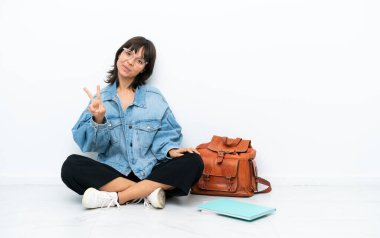 Genç bir kız öğrenci, beyaz arka planda tek başına oturuyor. Gülümsüyor ve zafer işareti gösteriyor.