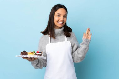 Pasta şefi, izole edilmiş mavi arkaplanda elinde büyük bir pasta tutarken mutlu bir ifadeyle selam veriyor..
