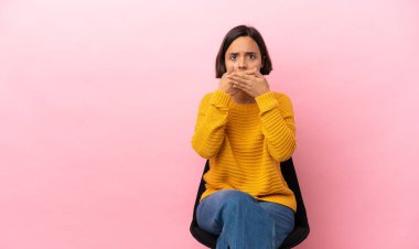 Genç melez bir kadın, pembe arka planda izole edilmiş bir sandalyede oturuyor. Ağzını elleriyle kapatıyor.