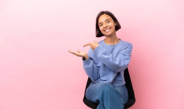 Genç melez bir kadın pembe arka planda izole edilmiş bir sandalyede oturuyor.