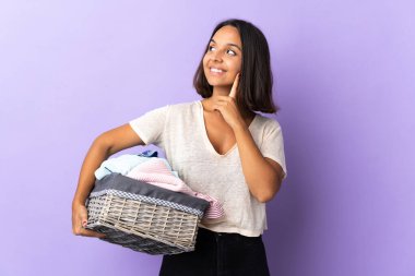 Mor arka planda izole edilmiş çamaşır sepetini tutan genç Latin kadın yukarı bakarken bir fikir düşünüyor.