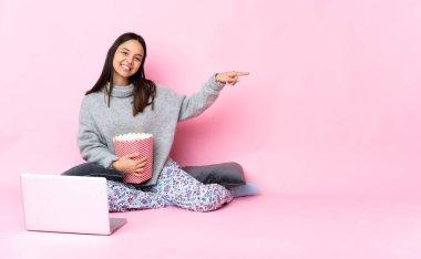 Genç melez bir kadın dizüstü bilgisayarında film izlerken patlamış mısır yiyor ve bir ürün sunuyor.