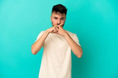 Mavi arka planda izole edilmiş genç beyaz bir adam sessizlik işareti gösteriyor.