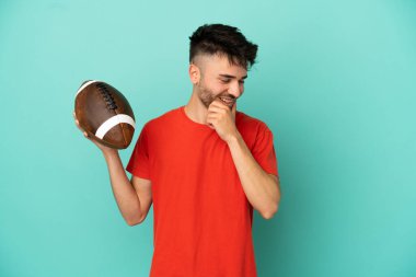 Genç beyaz adam ragbi oynuyor. Mavi arka planda izole edilmiş. Yan tarafa bakıyor ve gülümsüyor.