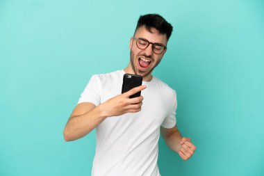 Genç beyaz adam mavi arka planda izole edilmiş cep telefonu kullanıyor ve zafer jesti yapıyor.