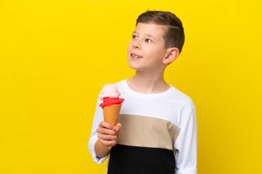 Sarı arka planda izole edilmiş kornet dondurması olan küçük beyaz çocuk yana bakıyor ve gülümsüyor.