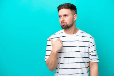 Young caucasian man isolated on blue background unhappy and pointing to the side