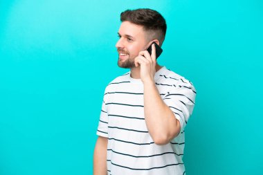 Kafkasyalı genç adam mavi arka planda izole edildi. Cep telefonuyla biriyle sohbet ediyor.