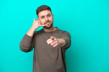 Kafkasyalı genç adam mavi arka planda izole edilmiş telefon hareketleri yapıyor ve ön tarafı işaret ediyor.