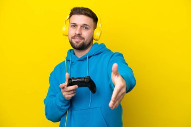 Sarı arka planda izole edilmiş bir bilgisayar oyunu kontrolörüyle oynayan beyaz bir adam iyi bir anlaşma için el sıkışıyor.