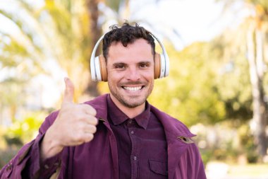 Genç beyaz adam açık havada müzik dinliyor ve başparmağını kaldırıyor.