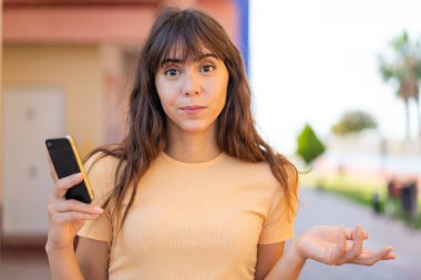 Dışarıda cep telefonu kullanan genç bir kadın omuzlarını kaldırırken şüphe hareketi yapıyor.