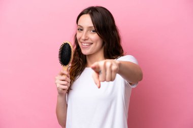 Pembe arka planda saç tarağı olan beyaz bir kadın kendine güvenen bir ifadeyle sana işaret ediyor.