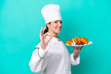Kafkasyalı genç aşçı kadın, mavi arka planda izole edilmiş waffle tutuyor. Parmaklarıyla işaret veriyor.