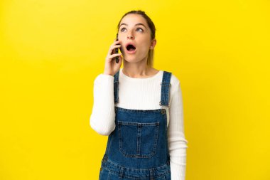 İzole edilmiş sarı arka plan üzerinde cep telefonu kullanan genç bir kadın yukarı bakıyor ve şaşırmış bir ifadeyle