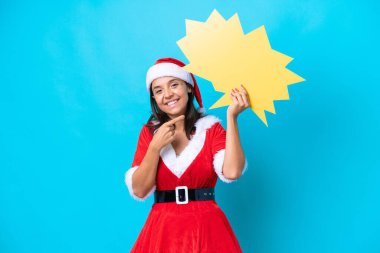 Anne Noel gibi giyinmiş genç İspanyol kadın mavi arka planda izole edilmiş boş bir konuşma balonu tutuyor ve ona doğrultuyor.