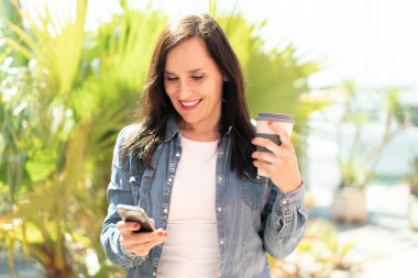 Orta yaşlı bir kadın cep telefonu kullanıyor ve elinde mutlu bir ifadeyle kahve tutuyor.