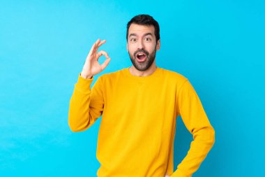 İzole edilmiş mavi arka plan üzerinde genç beyaz bir adam şaşırdı ve tamam işareti gösteriyor.