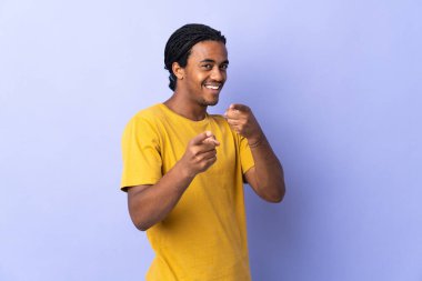Örgülü genç Afrikalı Amerikalı adam mor arka planda izole edilmiş şaşırmış ve ön tarafı işaret ediyor.