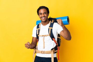 Sarı arka planda büyük bir sırt çantalı genç dağcı zaferi kutluyor.