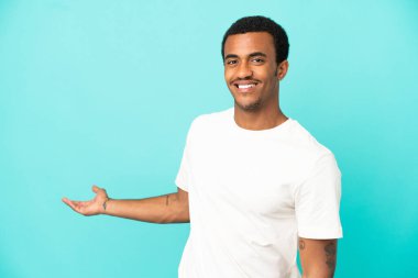 İzole edilmiş mavi arka planda oturan Afro-Amerikalı yakışıklı adam davet edildiği için ellerini yana uzatıyor.