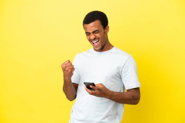 Afro-Amerikalı yakışıklı adam izole edilmiş sarı arka planda ve telefon zafer pozisyonunda.