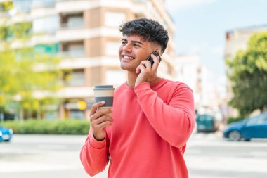 Dışarda cep telefonu kullanan ve elinde mutlu bir ifadeyle kahve tutan genç bir İspanyol.