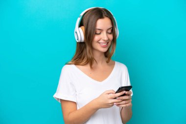 Genç beyaz kadın mavi arka planda izole edilmiş müzik dinliyor ve cep telefonu arıyor.
