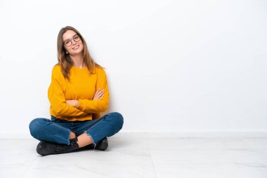 Beyaz arka planda, kolları çapraz ve ileriye bakan beyaz bir kadın yerde oturuyor.