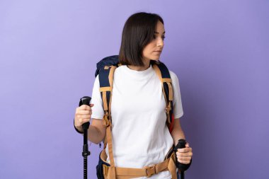 Sırt çantası ve yürüyüş direkleriyle mavi arka planda izole edilmiş beyaz bir kadın yan tarafa bakıyor.