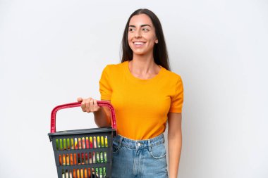 Beyaz arka planda izole edilmiş yiyecek dolu bir alışveriş sepeti tutan beyaz bir kadın yan tarafa bakıyor ve gülümsüyor.
