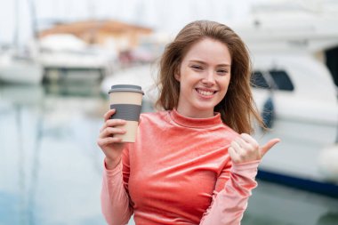 Young redhead woman holding a take away coffee at outdoors pointing to the side to present a product