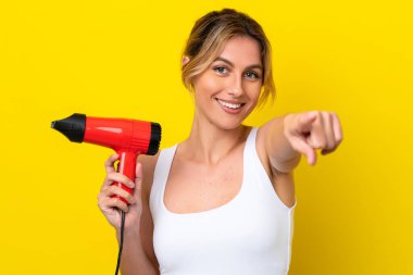 Genç Uruguaylı kadın elinde saç kurutma makinesiyle Don sarı arka planı izole ediyor. Kendine güvenen bir ifadeyle seni işaret ediyor.