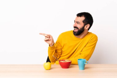 Masada kahvaltı yapan beyaz bir adam, yan tarafı işaret ediyor ve bir ürün sunuyor..